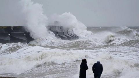 Two people stand in front of crashing waves 