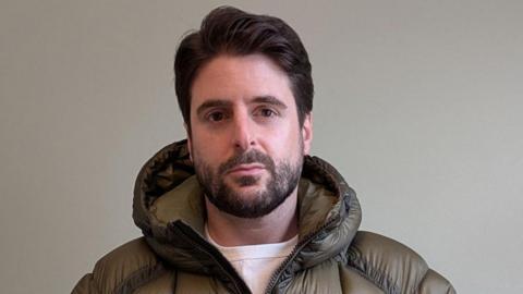 Alex Grossomanides has brown hair, brown eyes and a beard. He is wearing an olive green puffer coat and looking at the camera while standing in front of a white wall indoors.