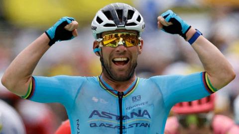 Sir Mark Cavenidsh, wearing a blue cycling top and shorts and a white helmet, holds his arms up and smiles