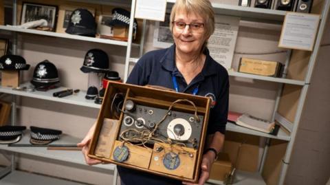 Sue Ward has short hair and glasses and is wearing a navy polo shirt and lanyard. She is holding a vintage looking radio with wires and boxes in a tan case. She is standing in front of shelves with exhibits such as police hats and helmets.
