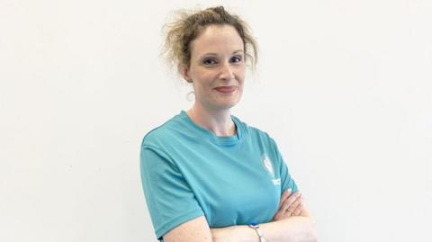 A woman in a blue top. Her arms are folded across her body. 