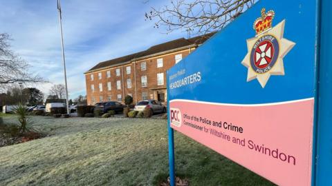 The view of the front of Wiltshire police HQ - the sign for it is close ot the camera, showing the force logo and that of the police and crime commissioner. The HW is a large red brick building with a flagpole in front. 