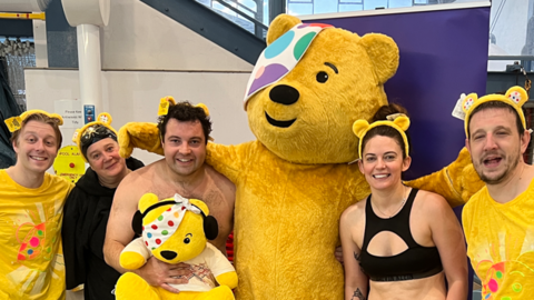 Five people wearing yellow Pudsey ear headbands smile and pose in a line with a lifesize Pudsey bear. George King is wearing a yellow Children in Need tshirt with an outline of Pudsey's face on the front, Cleah Heatherington is wearing a black swim cap and a back cover-up, Stuart Howells is holding a cuddly Pudsey to his bare chest, Zoie O' Brien is in black swim top and Isaac Chenery is wearing the same T-shirt as George 