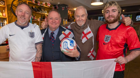Four men are standing together, with the man on each end of the line holding an England flag and wearing an England top. They are in a bar setting and the man second from the left is wearing a jacket, shirt and tie.