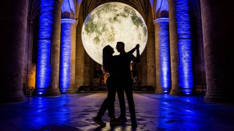 The silhouettes of a couple, a man and a woman who are both wearing glasses, are illuminated by a large, illuminated sculpture of the full moon inside Tewkesbury Abbey. The pillars have been lit up blue.