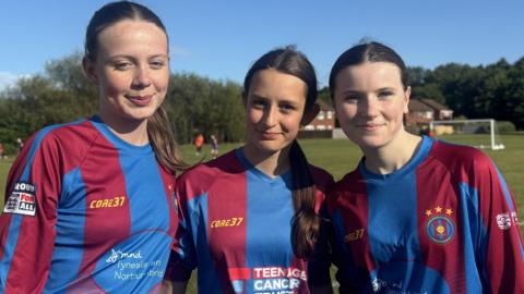 Three footballers stand together wearing blue and red striped football tops. They all have dark hair which is tied back and they are standing at the edge of a football pitch