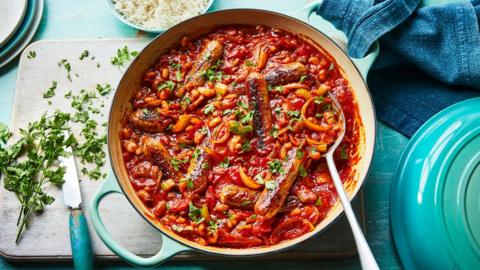 A wide oven dish filled with a spicy stew with sausages on top, sat on a wooden board with a blue cloth