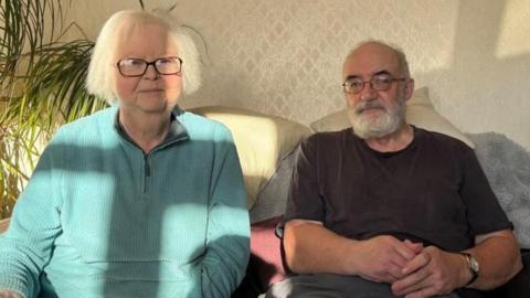 A woman in a light blue jumper sits next to her husband who is wearing a grey shirt, on a sofa. Behind them on one side are green fronds of a plant and behind them is beige patterned wallpaper