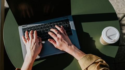 A woman's hands typing a laptop with a coffee. The laptop is situated on a green table.