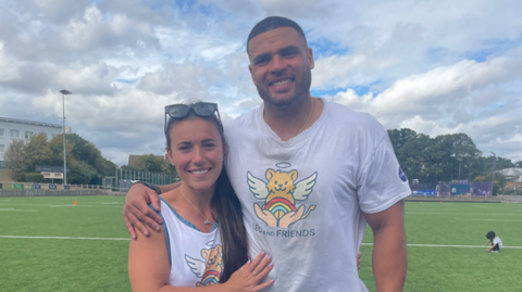 Zack George and his wife Samantha standing on a sports ground at Loughborough University. She has long dark hair and is wearing sunglasses on her head and a white vest. He is next to her, with cropped dark hair and wearing a white T-shirt. Both their tops are marked with a logo for Leo and Friends, which includes a cartoon lion portrayed with a halo and angel wings, sitting with a rainbow in outstretched human hands.   