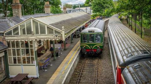 A train pulling into Bishop Auckland West station. A man sits at a table on the covered platform.