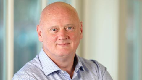 A bald man smiles at the camera. He wears a blue checked shirt. Blurred windows can be seen behind him.
