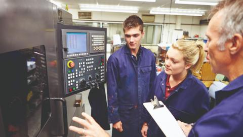 Two young people being shown a complicated piece of machinery by an older man. All three are wearing blue boiler-suits. A small screen with dozens of buttons is attached to the machine.