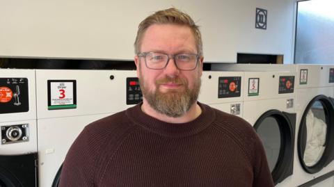 James Crumpton standing inside a launderette in front of a row of tumble dryer. He has light brown hair and a greying beard, and is wearing glasses and a dark brown jumper.