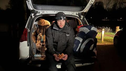 Emmet Meehan sits in the boot of a car during the challenge. He is wearing a black jacket with a headtorch. He is holding a half eaten slice of pizza.