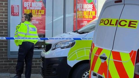 A female police officers stands outside a bookmakers. Two police vans are next to her and she is behind a police cordon. Red posters are in the shop window.