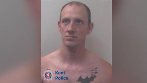 Sean Boyle - a close up police mugshot of a man naked from the top up with a large bat tattoo on his left breast. He is looking seriously to the camera's right