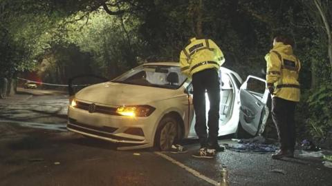 Two police officers in yellow high-vis jackets inspect a white car that is crashed at the side of the road. 