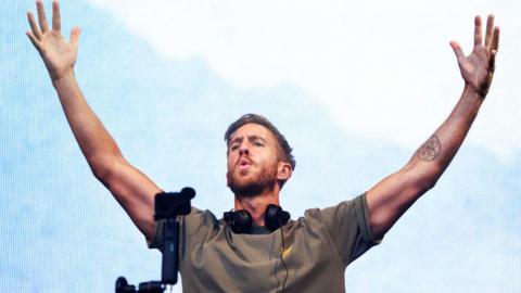 Calvin Harris stands behind a mixing desk with headphones around his neck. He has his arms raised in the air