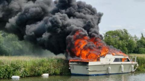 A white boat on fire with black smoking billowing from it. Beyond it is a river bank, a field, and trees in the background. 