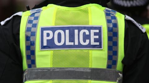 A police officer wearing a hi-viz vest over a black shirt. The word POLICE is in capital letters in white writing on a blue background on the back of the vest.