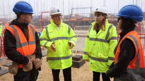 Keir Starmer and Angela Rayner wear yellow high visibility coats and white hard hats. To their left and right are 2 people wearing orange high visibility jackets and blue hard hats. 