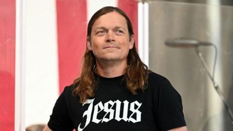 Brad Arnold of the rock band 3 Doors Down smiles . He has long hair and black T-shirt with 'Jesus' written across the front. 
