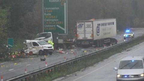 The scene of the collision on the A30. There is a highways maintenance van which has its rear end destroyed. A highways maintenance small lorry is near it and there is a HGV which reads 'Conway Bailey Transport' on the side. There is a stationary police car with flashing lights behind that lorry. Debris covers the carriageway. A road sign reads Bodmin A30 straight on and left turning to Launceston, Bude, Callington and Liskeard.