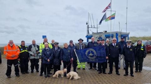 Several members of National Coastwatch Mablethorpe and others standing with the blue National Coastwatch Institution flag next to the station in Mablethorpe