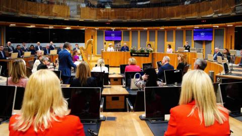 A general view of the Senedd during a royal visit, with the King and Queen Camilla sat in the centre, and a harp in view. Vaughan Gething, who was first minister at the time, is stood with  his back to the camera.