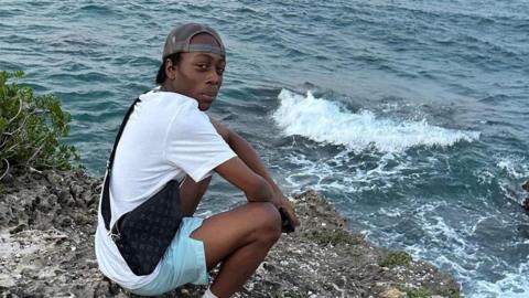 Deonte Mowatt-Slater in a holiday photo, crouching near the sea. He wears shorts, T-shirt and a backwards cap, and a YSL branded crossbody man bag. He is looking at the camera.