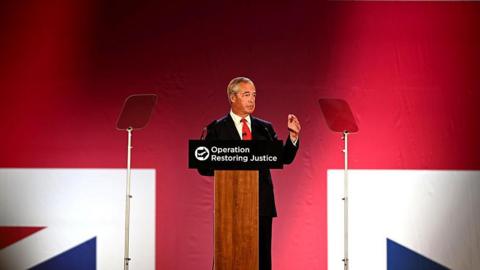 Nigel Farage, wearing a black suit and a red tie, standing at a podium that says "Operation Restoring Justice" on it. He's talking into a microphone, with a large Union Jack projected onto the wall behind him.