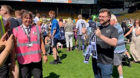A fan posing with the Port Vale promotion trophy. Hundreds of fans on a football pitch. They are on green grass and there is a stand behind them. 