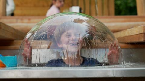 A boy pokes his head up in a glass lid, which has water dripping down the outside.