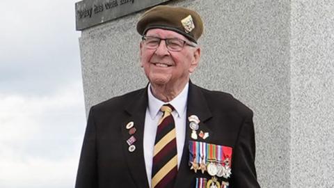 Richard Brock smiling in a white shirt and black suit jacket with his regimental cap, tie and medals.