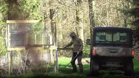 A person in camouflage clothing stands beside a wooden enclosure with wire mesh in a sunlit forest clearing. They appear to be opening or closing a small gate. A dark utility vehicle is parked on a grassy track to the right, and tall trees with sparse foliage fill the background.