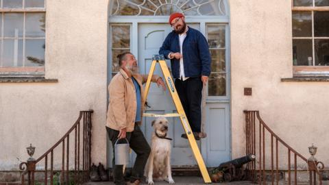 This image shows two adults standing at the entrance of a house, posed around a yellow step ladder, with a dog sitting between them.