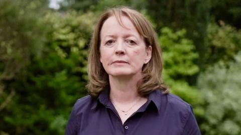 Dr Susan Gilby looks at the camera. She is wearing a purple blouse and a necklace. She has shoulder length light brown hair and there is greenery blurred out in the background.