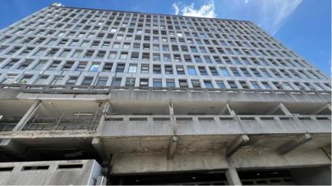 Terminus House is a grey building, with 14 storeys, with hundreds of windows. There is a blue sky above.
