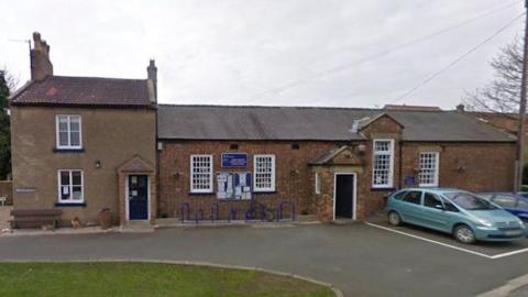 A small, one-storey red-brick school with a black door and white windows.  There is a grey extension, which has two floors.  Two cars are parked outside and the sky above is grey.