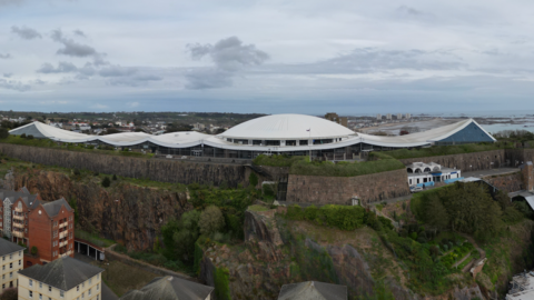 A wide panoramic view of a hilltop complex featuring Fort Regent - a large, white, curved-roof building at the centre, surrounded by stone walls and green embankments.