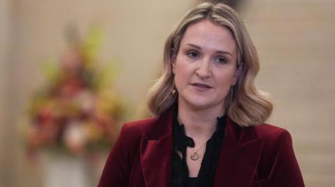 Helen McEntee is looking away from the camera. She has shoulder length blonde hair. She is wearing a red jacket and black blouse.