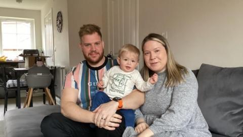 Emma and Daniel sit on a dark grey sofa with toddler Vinnie on their lap and the kitchen and dining table in the background behind them. They are all looking at the camera and smiling.