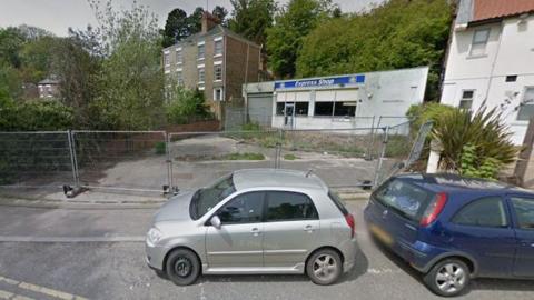 Two cars parked by a fenced-off area of concrete. A building is behind the former forecourt with "Express Shop" written on it.