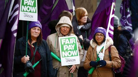 Three women wearing purple hats hold fair pay now placards and purple flags at a street rally.