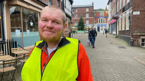 A man with cropped fair hair and stubble stands on a quiet tiled street with a few pedestrians in the background. He is wearing an orange coat covered by a yellow hi-vis vest. A cafe and outside tables are to the left of the image. A pub, shop fronts and a Christmas tree can be seen in the distance.