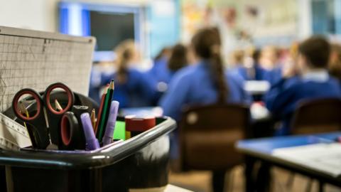 A blurred background shows pupils in school uniforms facing a school blackboard, in the forefront is a pot of school stationary