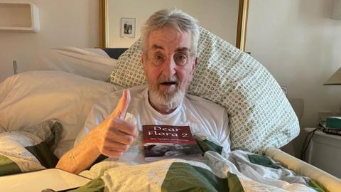 A man with short grey hair and a beard, is wearing a white T-shirt. He is sitting up in a bed, with a pillow behind him, and is looking at the camera and giving a thumb's up with his right hand. On his chest is a book called "Dear Flora 2".