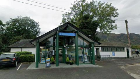 Google Maps image of Coniston Visitor Information Centre. It has a slanted roof, like an upturned V, and glass frontage. Trees and hills can be seen in the background.