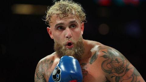 Jake Paul in action during a fight, he's shirtless and wearing blue boxing gloves.